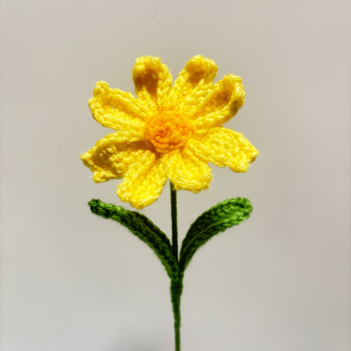 Yellow crocheted flower with a green stem on a light gray background.