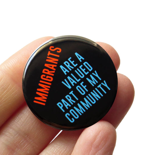 Black button with red and blue ‘Immigrants, Are a valued part of my community’ text held by a hand on a white background. 
