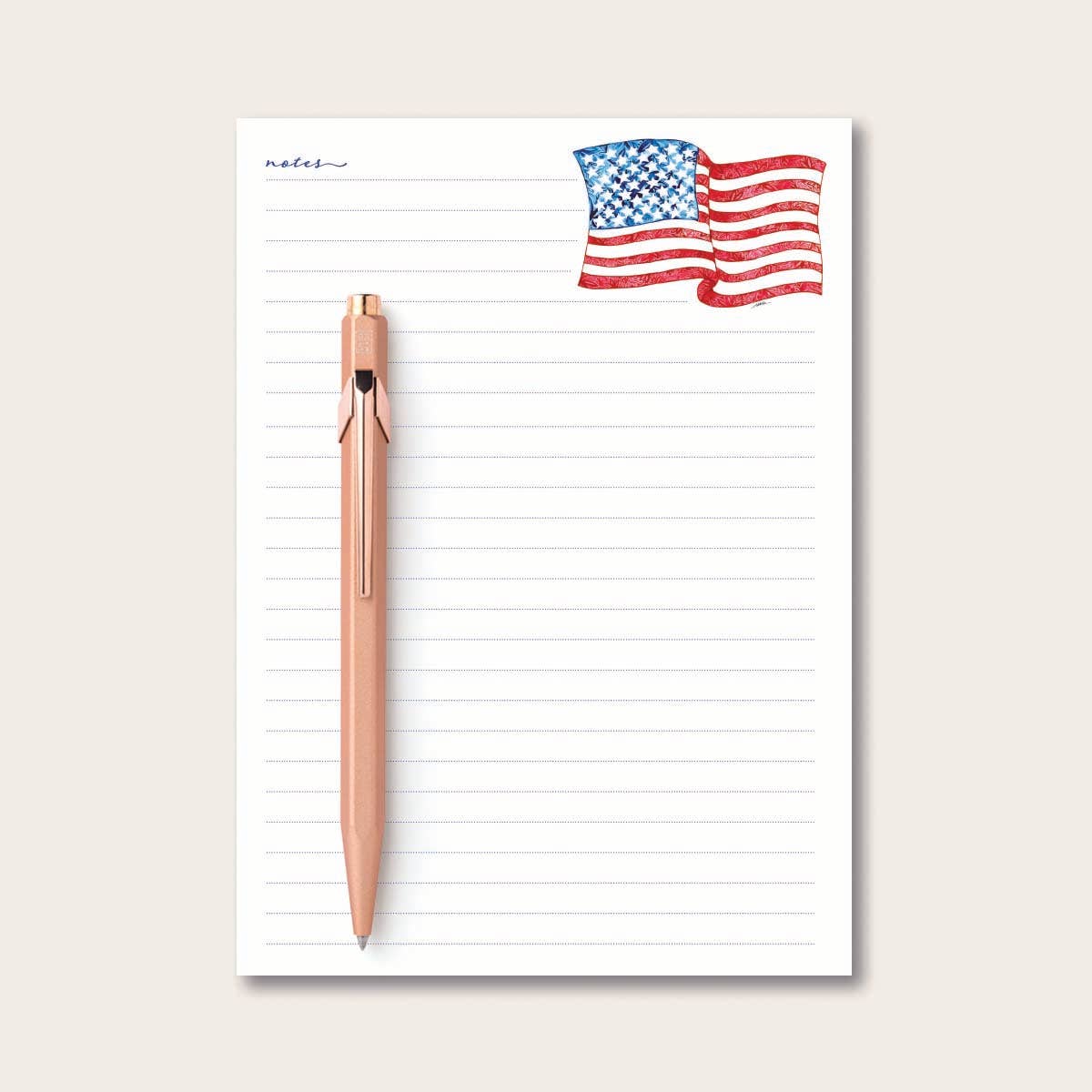 Notepad with American flag design at the top and a rose gold pen on a light beige background