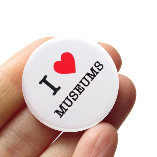 White button badge with ‘I heart museums’ text held in a hand against white background.