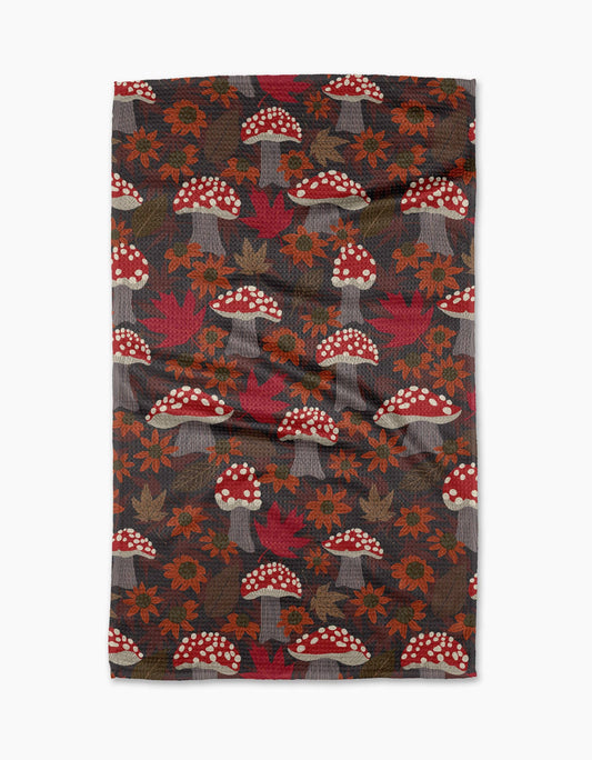 A decorative dark brown towel with fall colored leaves and flowers next to various red mushrooms with white spots on a white background. 