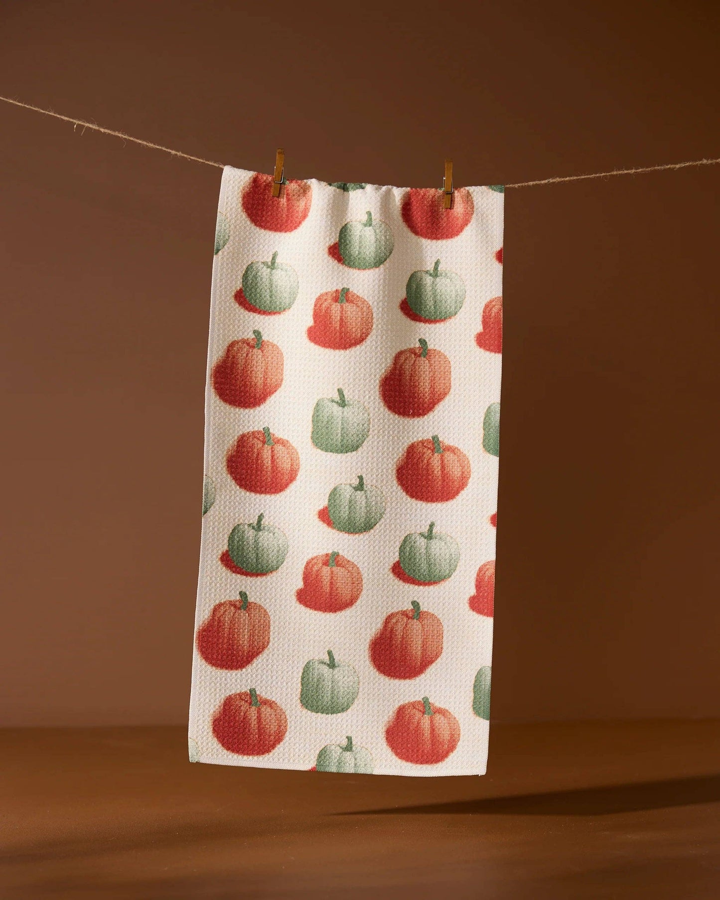 A decorative white tea towel with orange and green pumpkins. Hanging from a line against a brown background. 