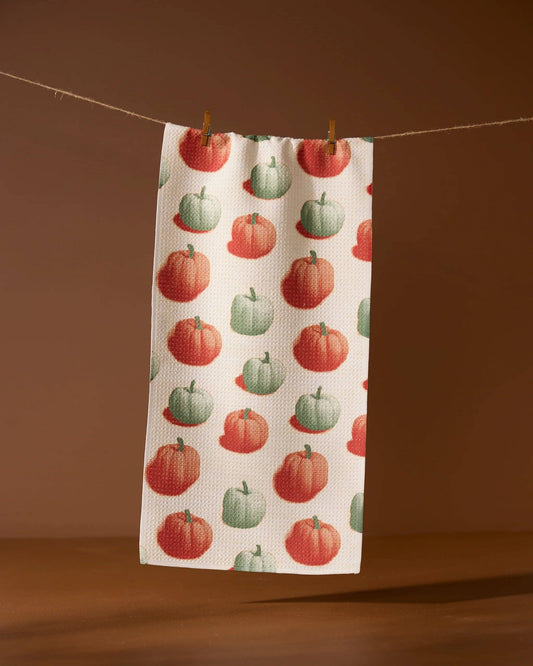 A decorative white tea towel with orange and green pumpkins. Hanging from a line against a brown background.