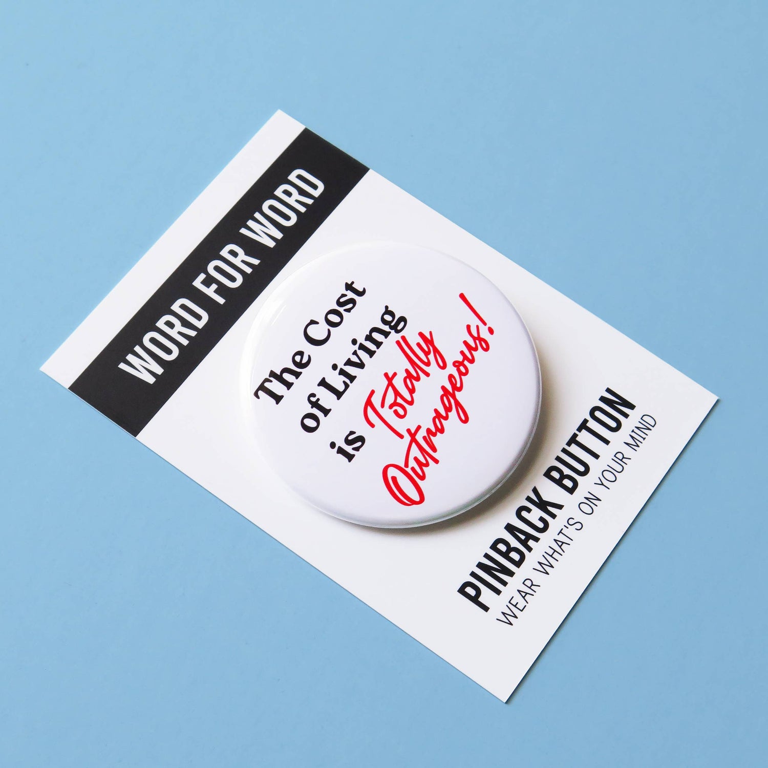 White button with text 'The Cost of Living is Totally Outrageous!' On black and white packaging ‘Word for word. Pinback button wear what’s on your mind’ packaging. On a bright blue background. 
