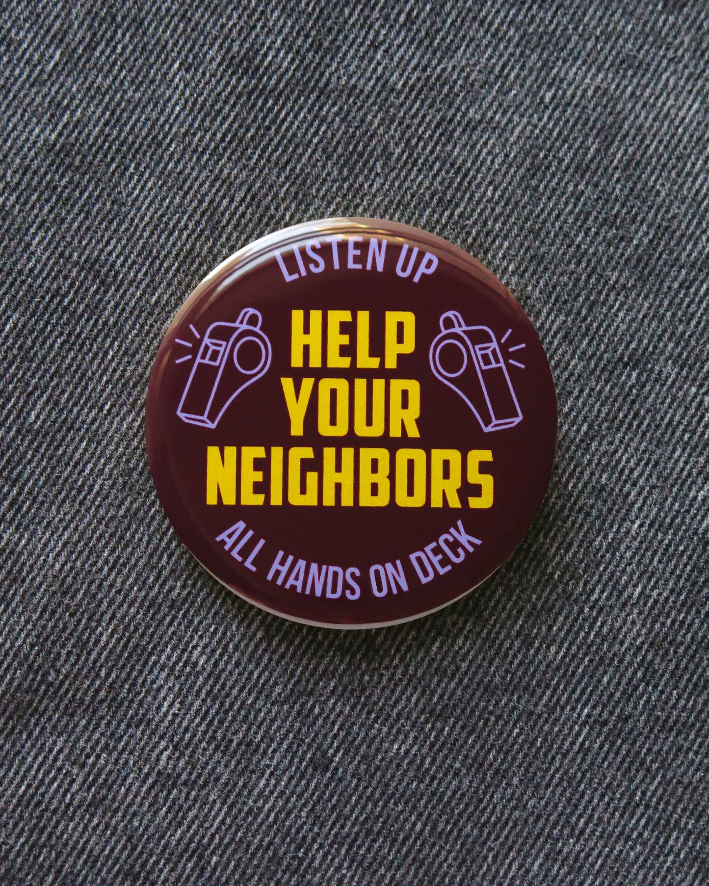 Brown button with ‘Listen up, Help your neighbors, all hands on deck’ and purple whistle decor on a denim pattern background. 