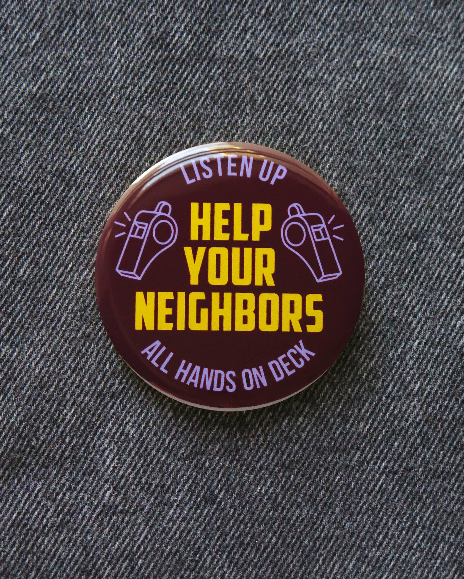 Brown button with ‘Listen up, Help your neighbors, all hands on deck’ and purple whistle decor on a denim pattern background. 