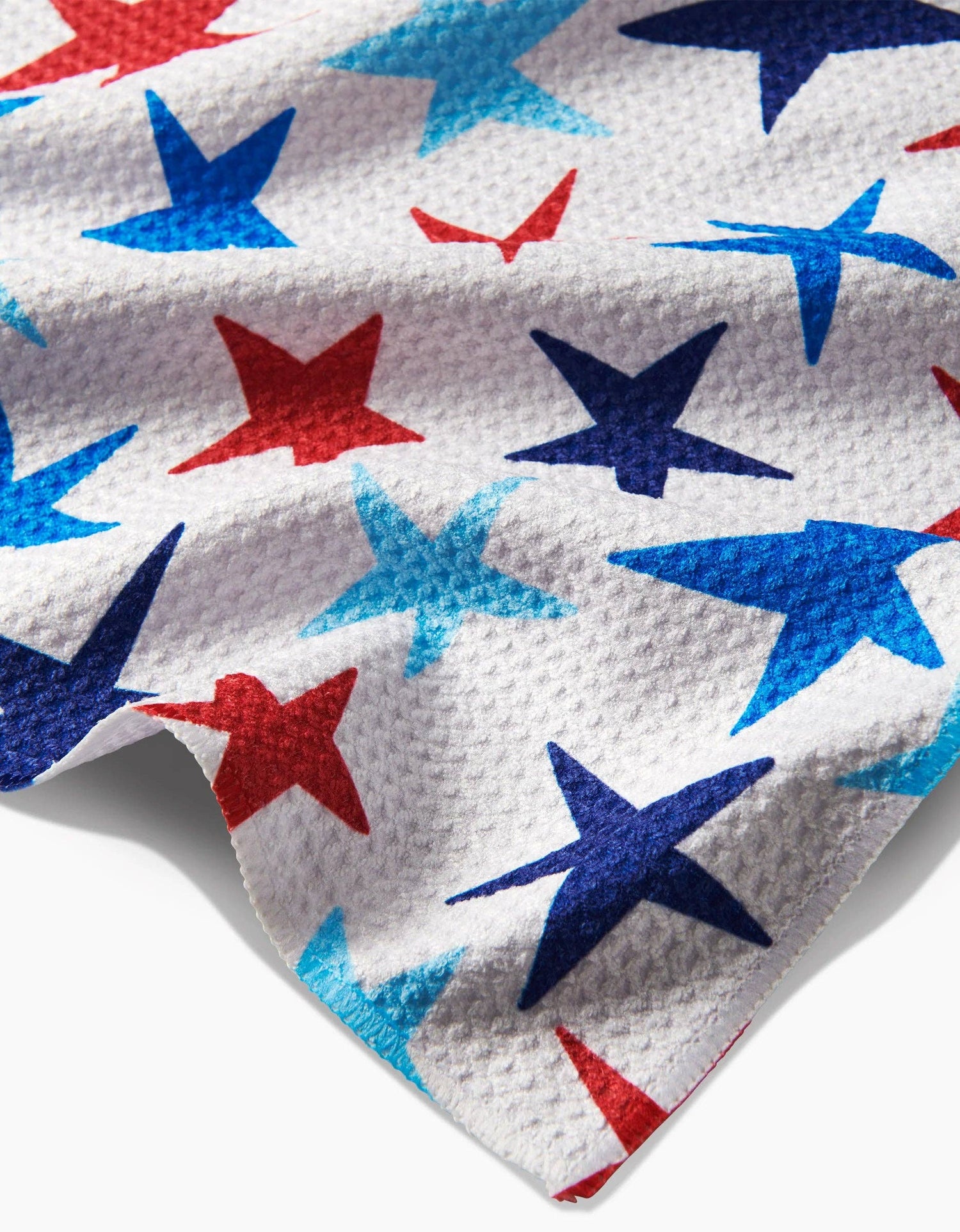 Corner close up of a decorative white tea towel with red, dark blue, and light blue stars of various sizes on a white background. 