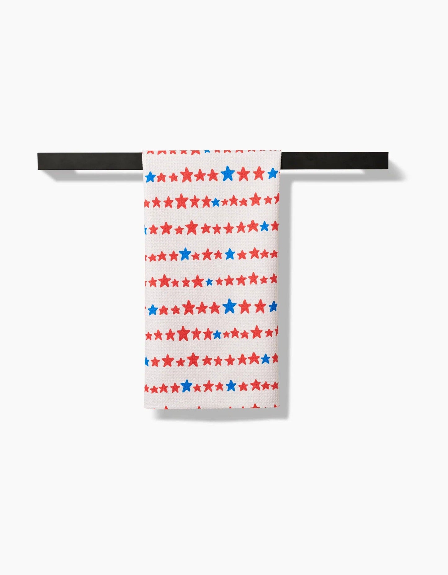 A decorative white tea towel with red and blue stars in a stripe like pattern hanging from a towel rod on a white background.