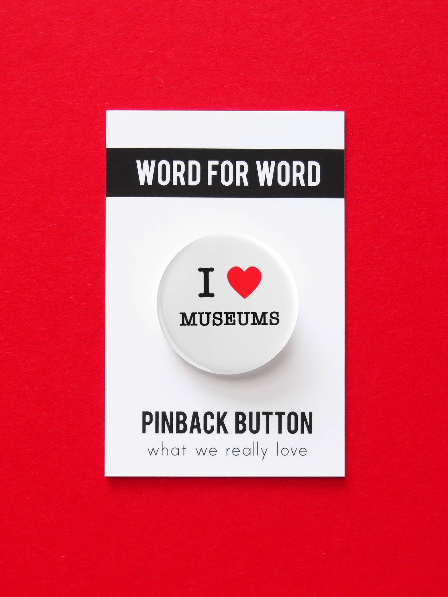 White button with ‘I heart museums” text on black and white ‘Word for word, Pinback button what we really love’ packaging. On a red background.