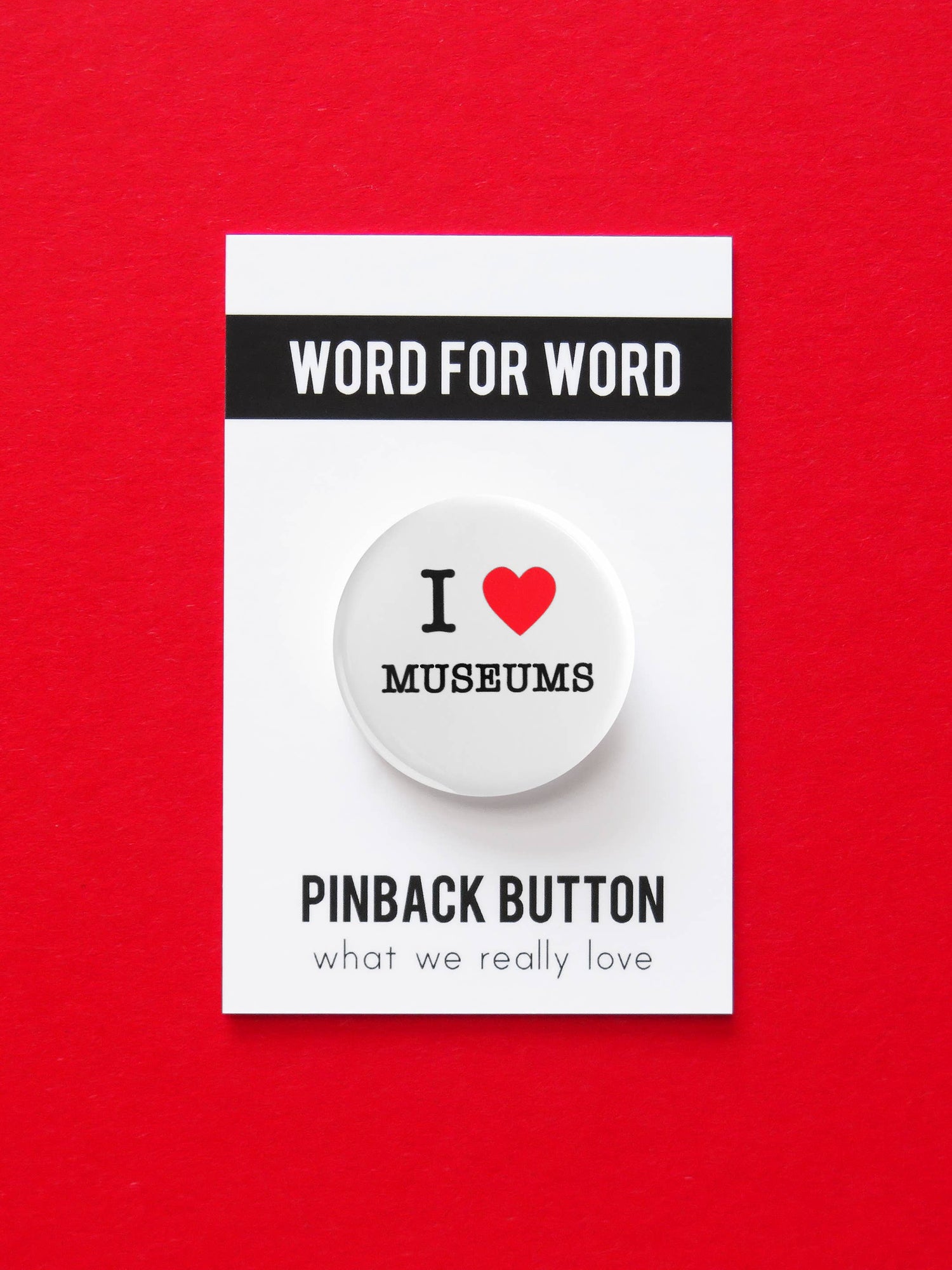 White button with ‘I heart museums” text on black and white ‘Word for word, Pinback button what we really love’ packaging. On a red background.