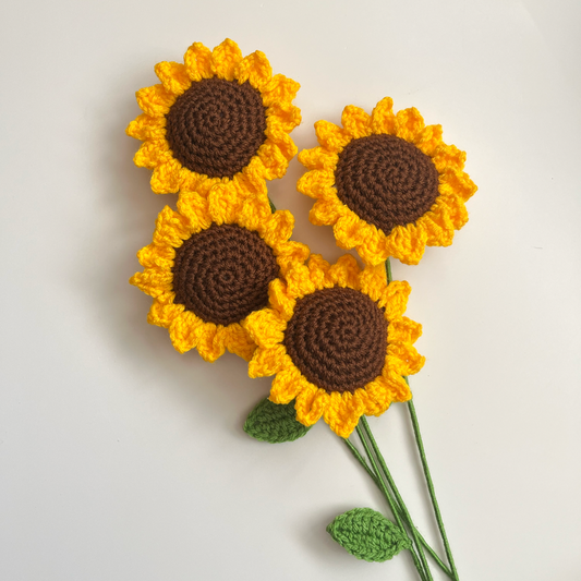A crochet representation of sunflowers with yellow petals and brown centers, with green leaves, against a white background.
