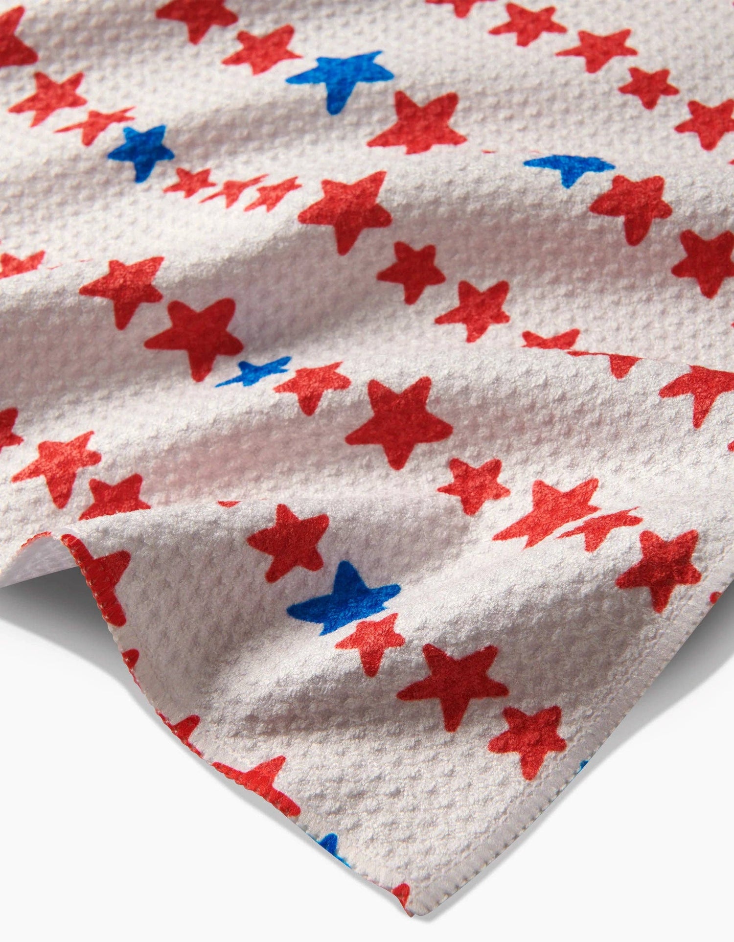 Corner close up of decorative white tea towel with red and blue star arranged in a stripe pattern on a white background. 