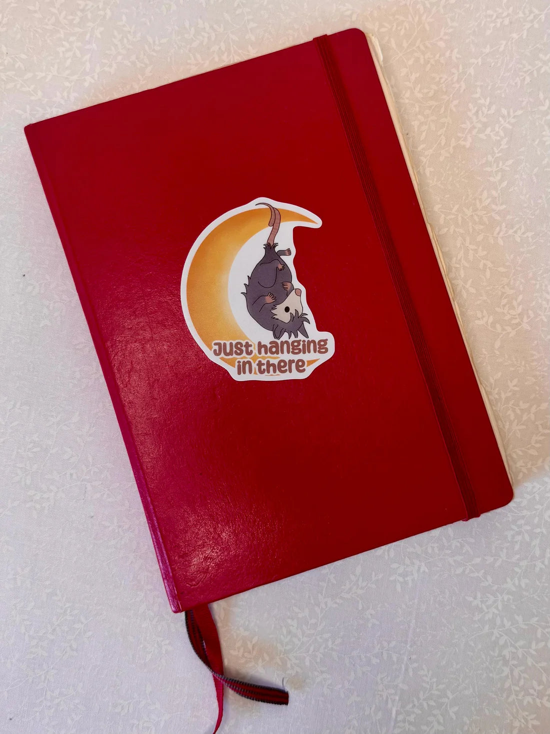 Red notebook with a sticker of an opossum on a crescent moon and text 'just hanging in there' on a light background