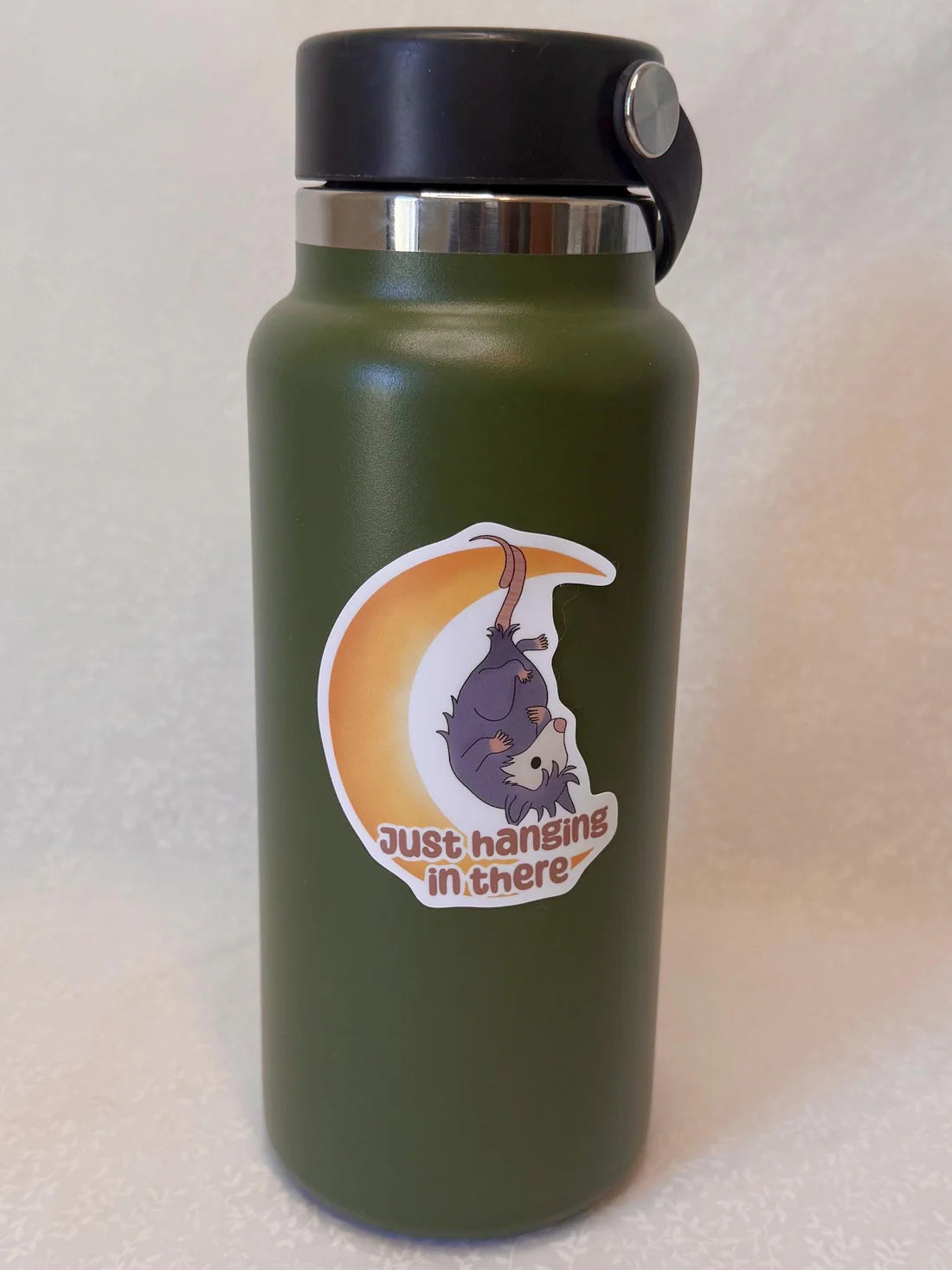 Green water bottle with a sticker of an opossum on a crescent moon and text 'Just hanging in there.'