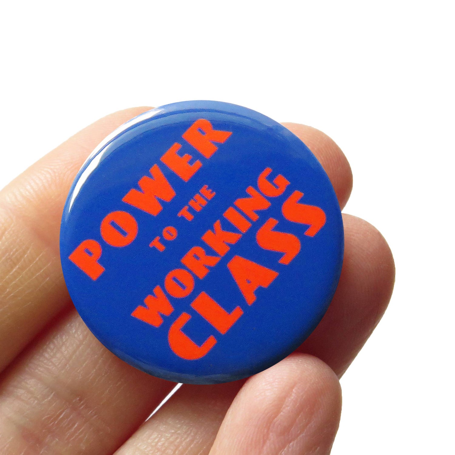 Blue button with orange text 'Power to the Working Class' held in a hand against a white background.