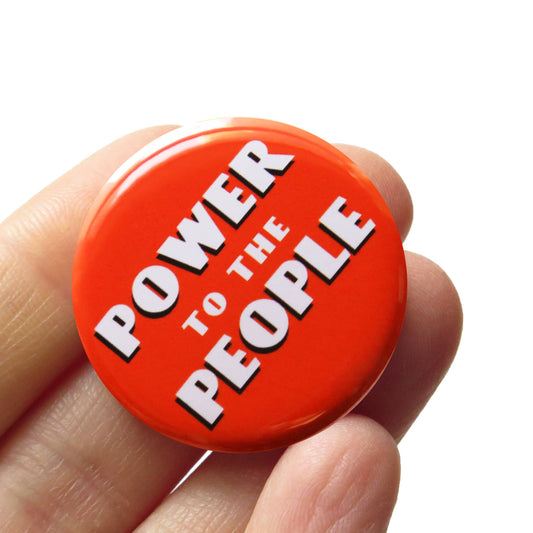 Red button with white text stating 'Power to the People' held in a hand against a white background