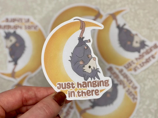 Sticker with an opossum on a crescent moon and 'Just hanging in there' text, held by a hand.