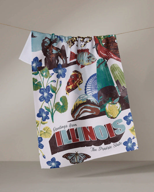 A decorative tea towel with ‘Greeting’s from Illinois the prairie state’ text. Decor on the towel includes flowers, bugs, food, and landmarks. On a beige background hanging from a line.