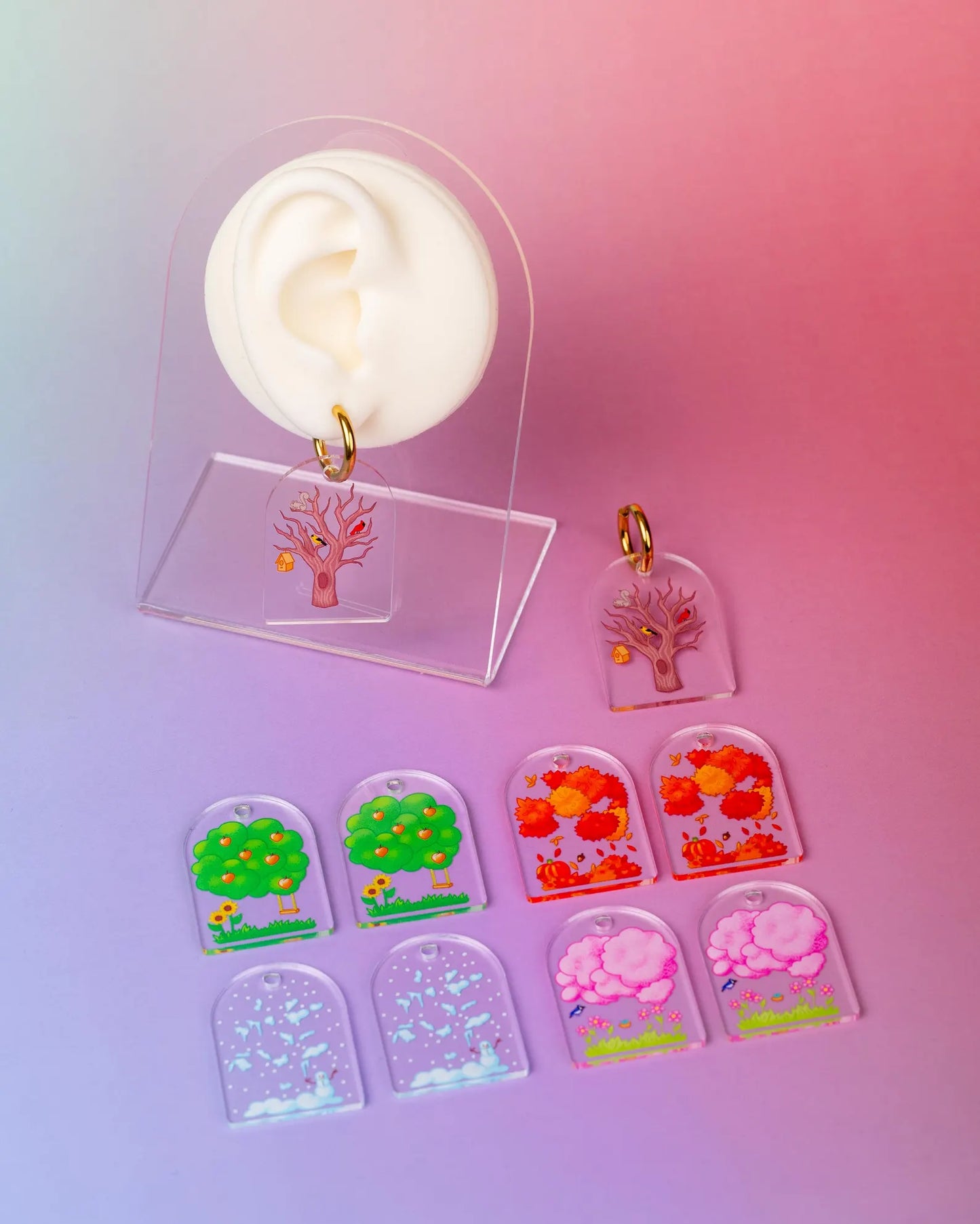 Set of interchangeable earrings depicting the four seasons through trees displayed on a fake ear. On a colorful blue and pink background. 