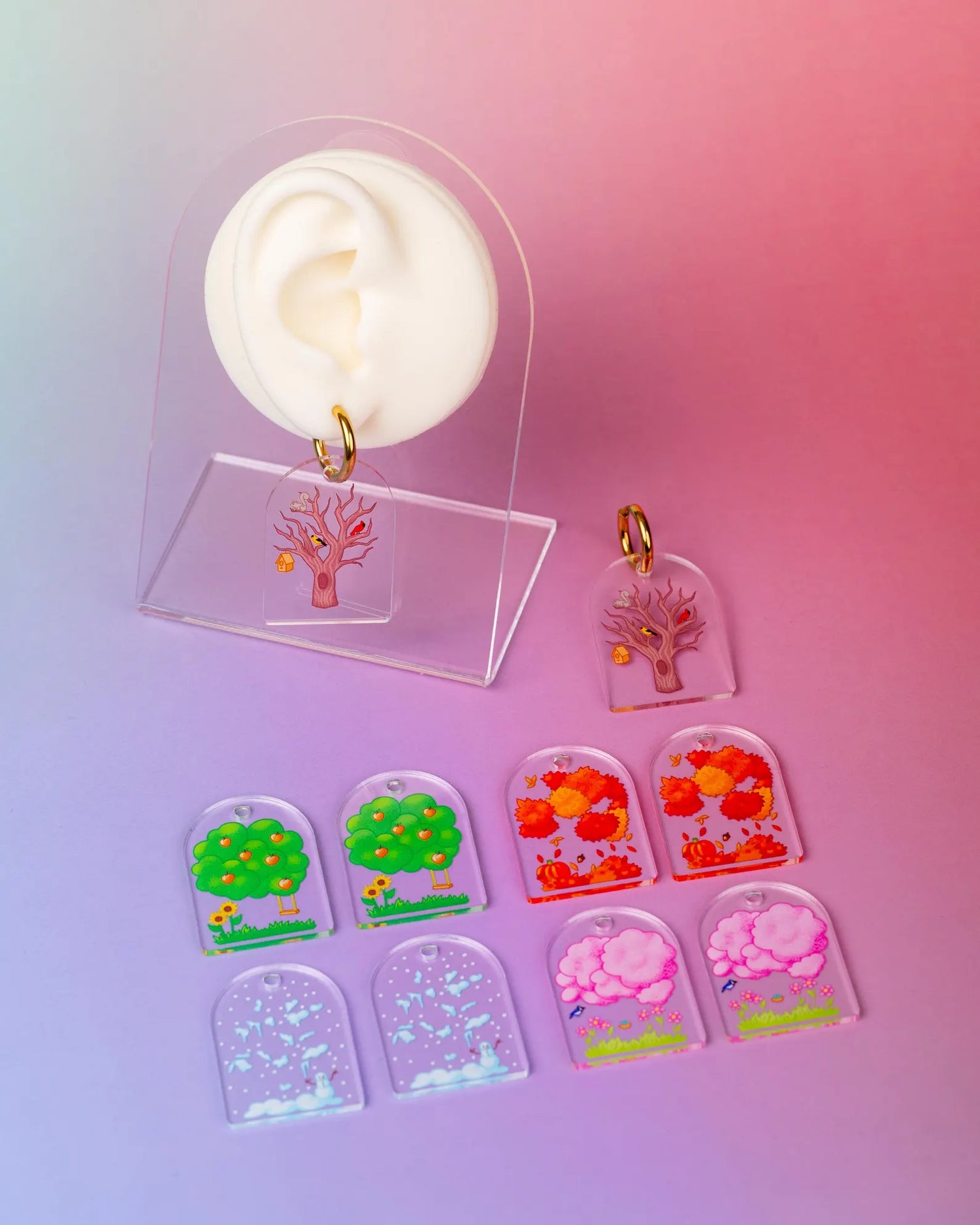 Set of interchangeable earrings depicting the four seasons through trees displayed on a fake ear. On a colorful blue and pink background. 