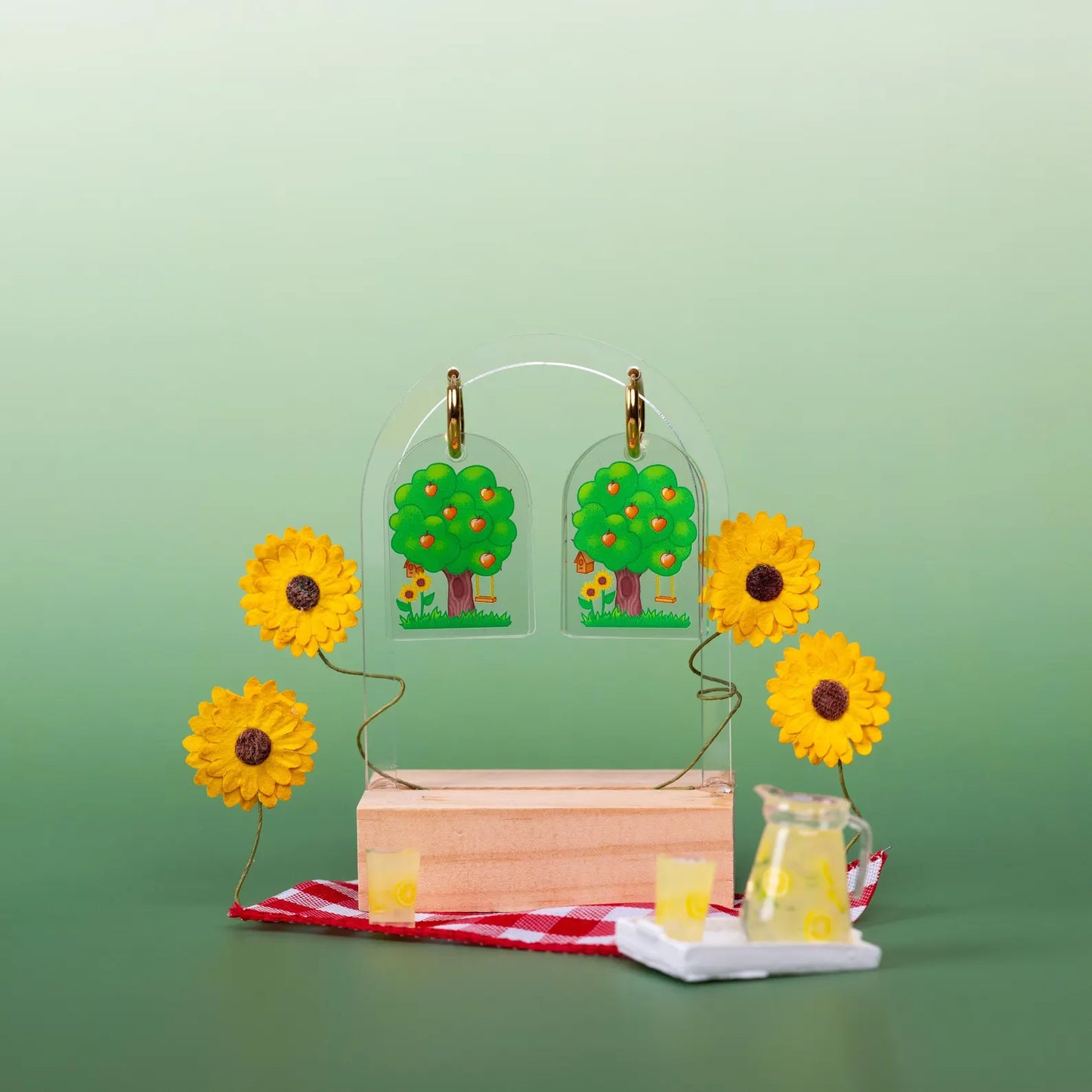 Set of summer earrings designed with a tree full of apples. Displayed on a wooden block on a small picnic blanket with fake sunflowers and lemonade. On a green and light green background. 