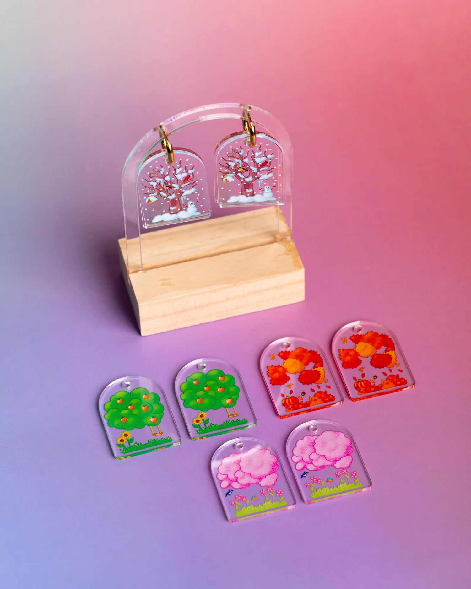 Set of interchangeable earrings with the four seasons winter, summer, fall, and spring depicted through trees. On a colorful blue and pink background. 