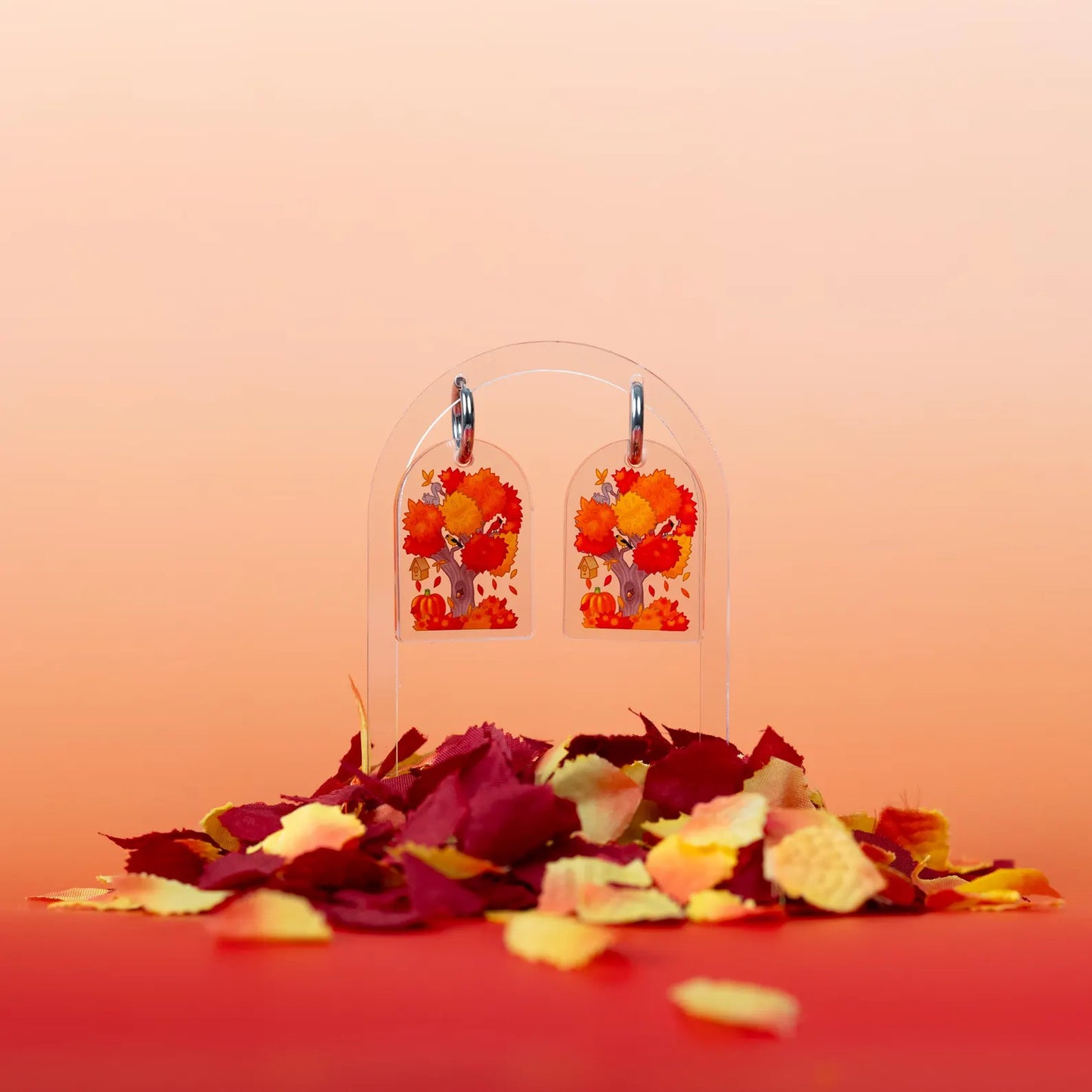 Set of the fall design earrings showing bright red, orange, and yellow leaves. Displayed above a pile of fake fall leaves and on an orange and light orange background. 