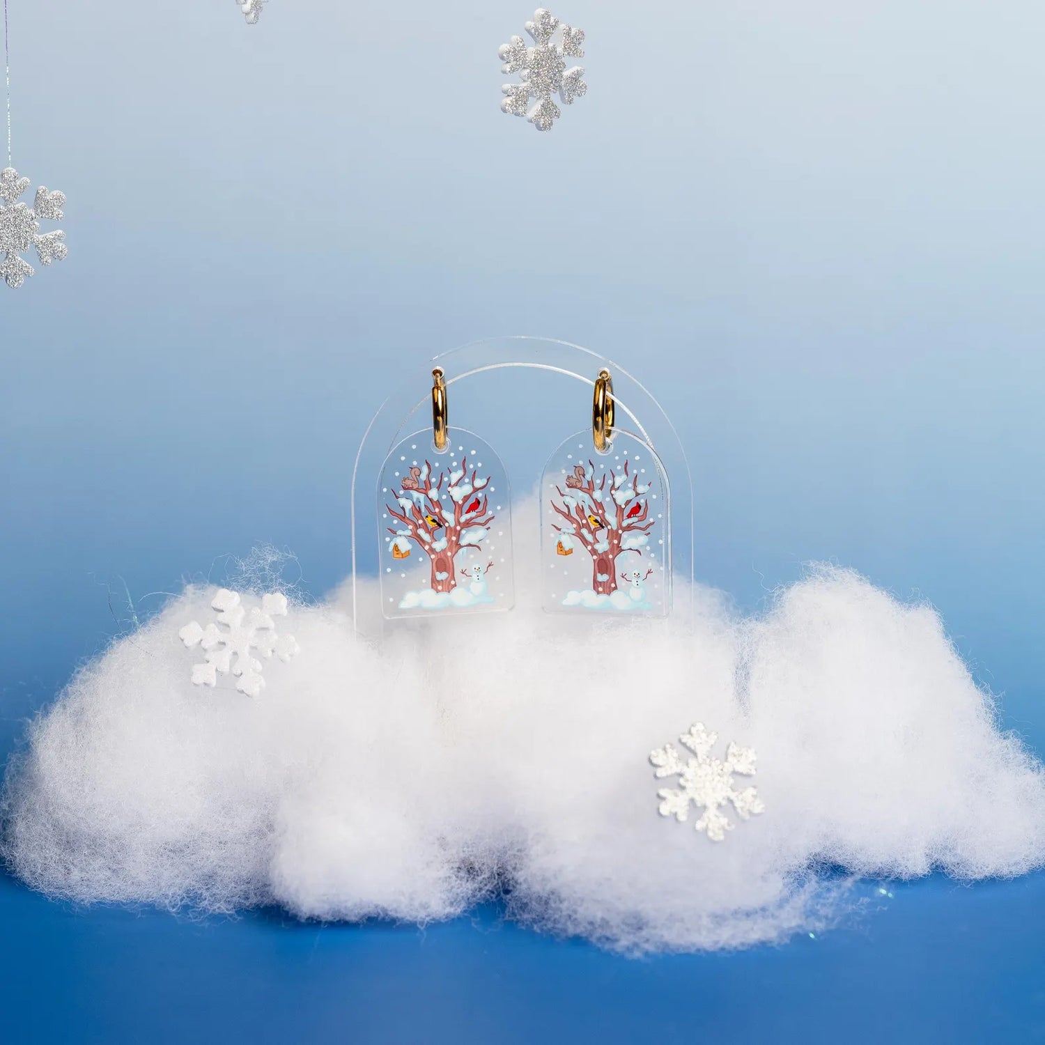 Set of winter themed earrings shown through a snow covered tree. Displayed above fake snow and snowflake decor on a blue and light blue background. 